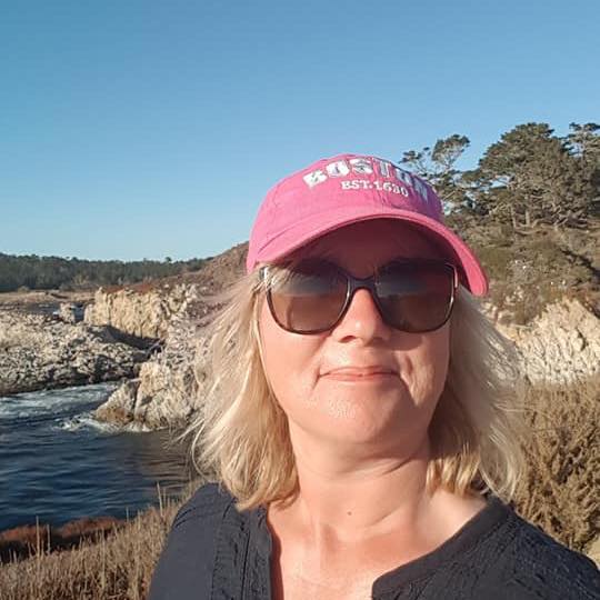 Jane Seale smiling outdoors, with the sea and rocky cliffs behind her.